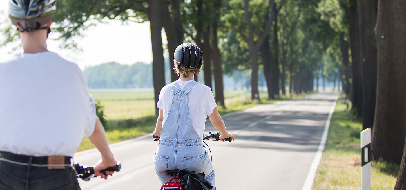 Zwei Personen mit Fahrradhelmen fahren auf einer schmalen Landstraße ohne separaten Radweg.